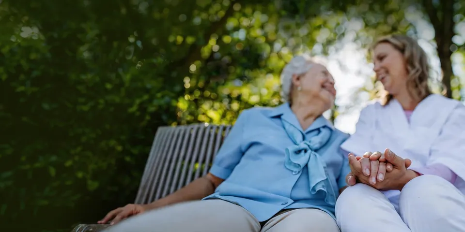 An older woman and a caregiver holding hands on a garden bench